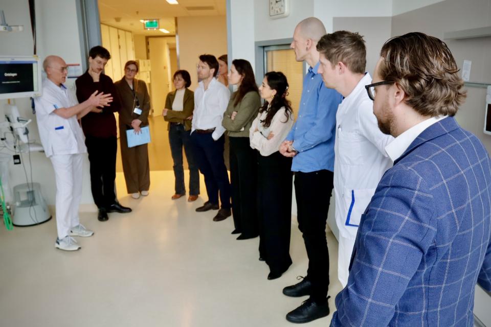 Leden en stafleden van de parlementaire enquêtecommissie Corona staan in een ziekenhuiskamer. Een arts spreekt hen toe.