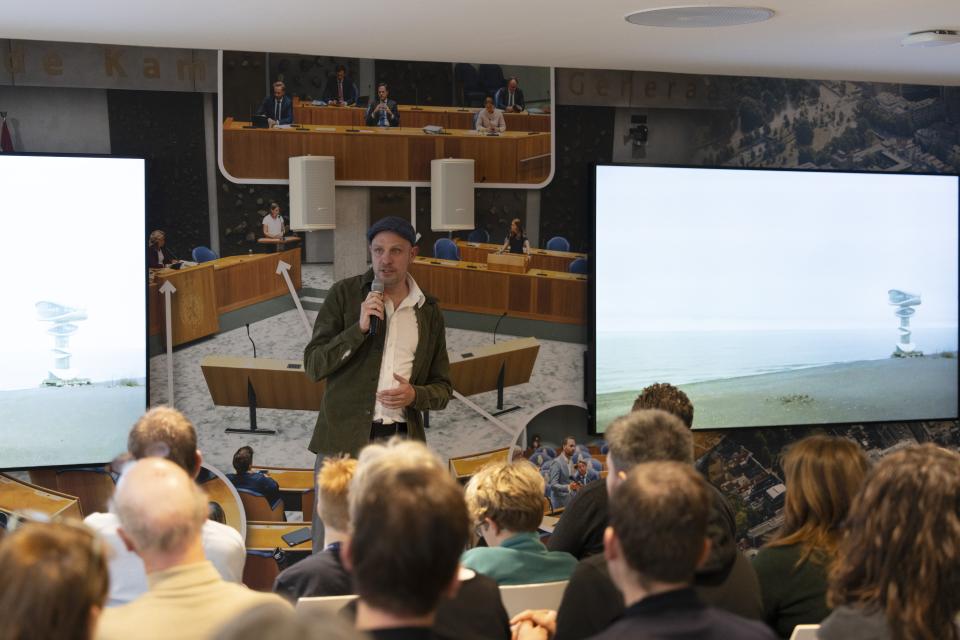 Jelle Brandt Corstius staat voor een publiek in de Filmzaal van de Tweede Kamer.