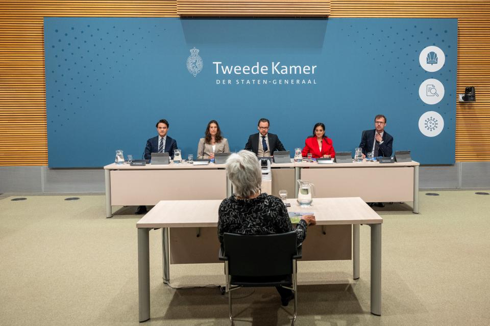 De leden van de parlementaire enquêtecommissie Corona zitten naast elkaar aan een lange tafel in de Enquêtezaal. Tegenover hen zit de verhoorde, Marion Koopmans.