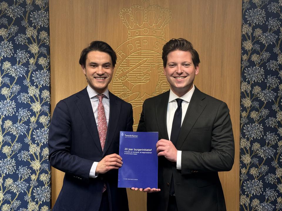 Twee personen staan voor een gebloemde wand en houden samen een donkerblauw document vast. 