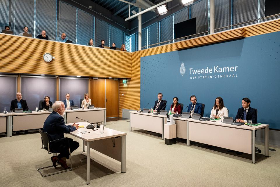 De leden van de parlementaire enquêtecommissie zitten naast elkaar in de Enquêtezaal. Tegenover hen zit Jaap van Dissel.
