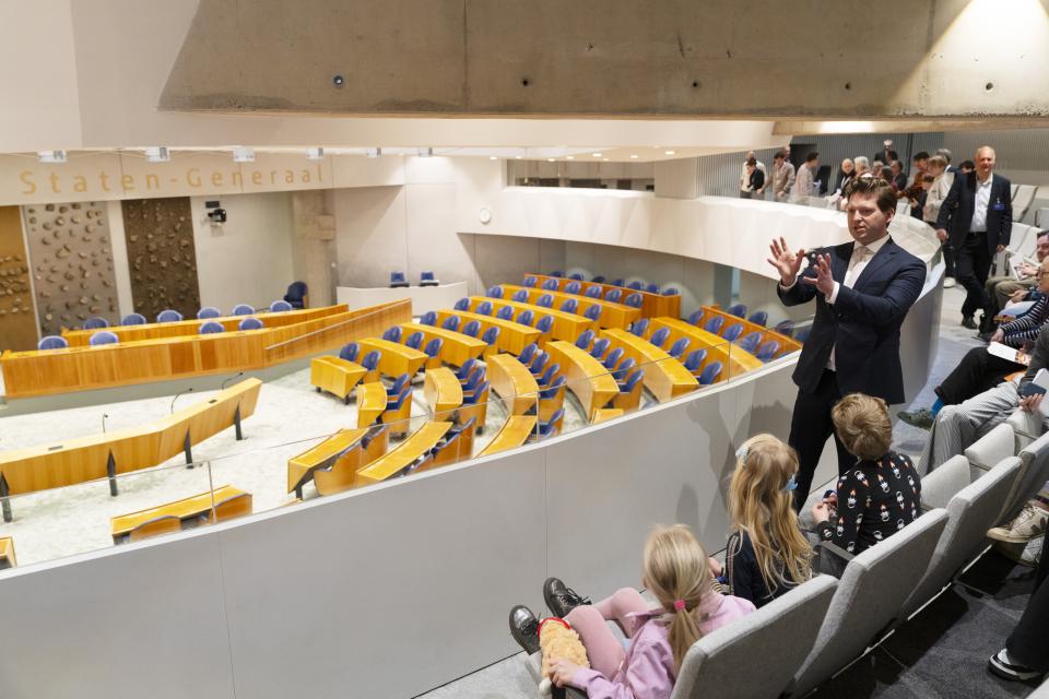 Kamervoorzitter Thom van Campen staat op de publieke tribune van de plenaire zaal. Hij vertelt een groep kinderen over het werk van de Tweede Kamer.