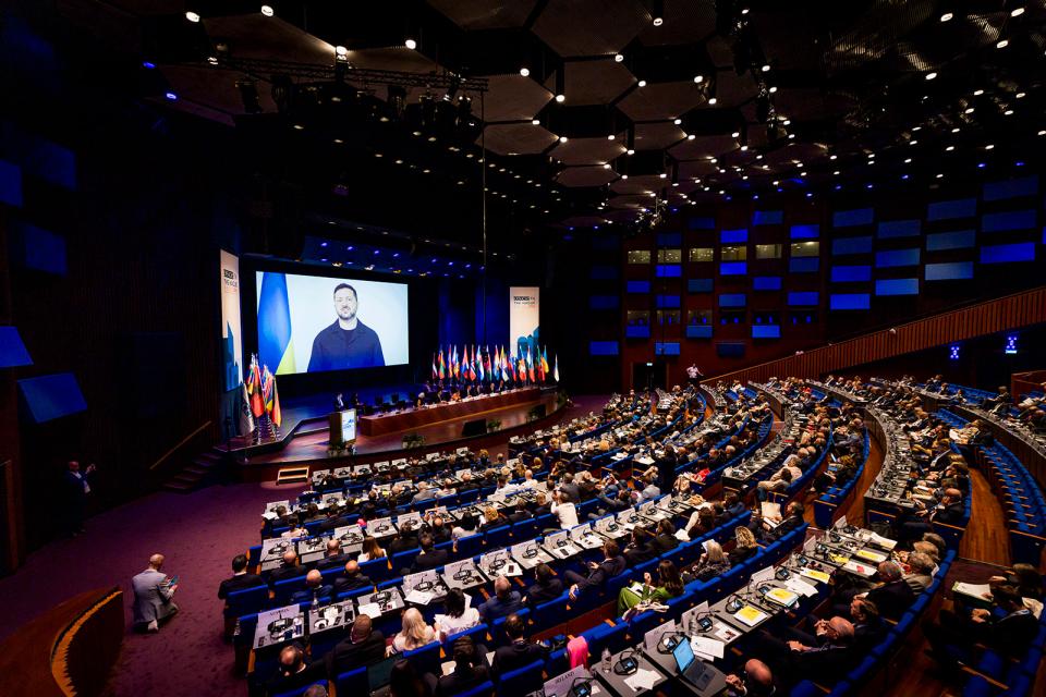 Plenaire zaal in het World Forum. Aanwezigen kijken naar een groot scherm met daarop een videotoespraak van de Oekraïense president Volodymyr Zelensky.