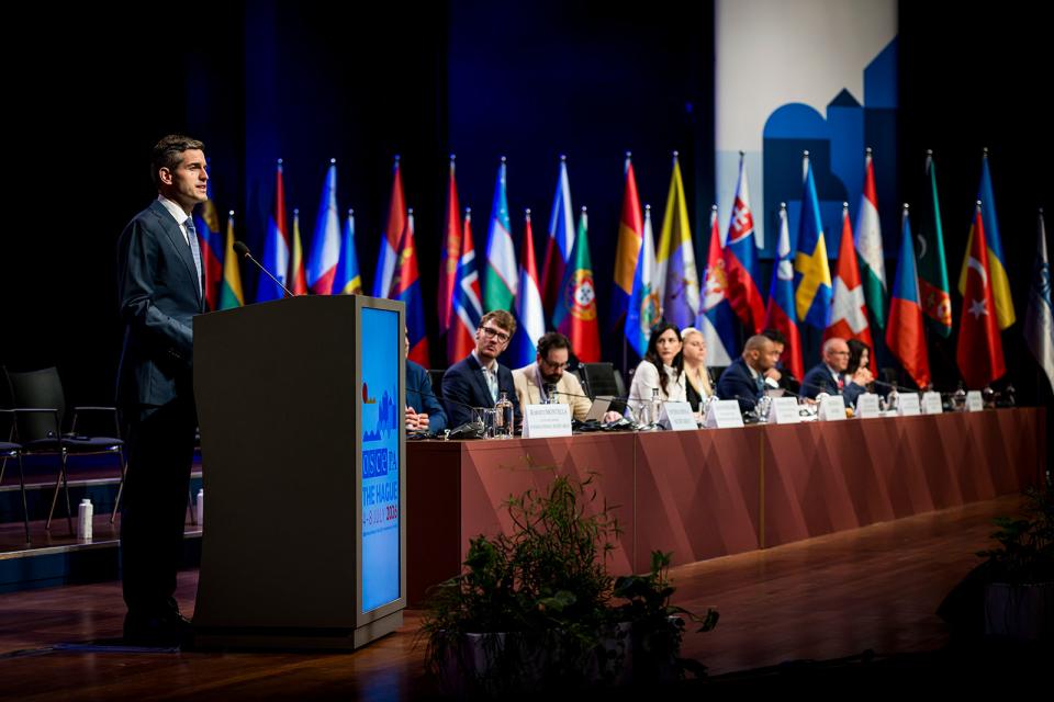 Minister van Buitenlandse Zaken Tom Berendsen staat achter een katheder en spreekt de zaal toe vanaf een podium. Achter hem een aantal personen aan een tafel en een rij vlaggen.