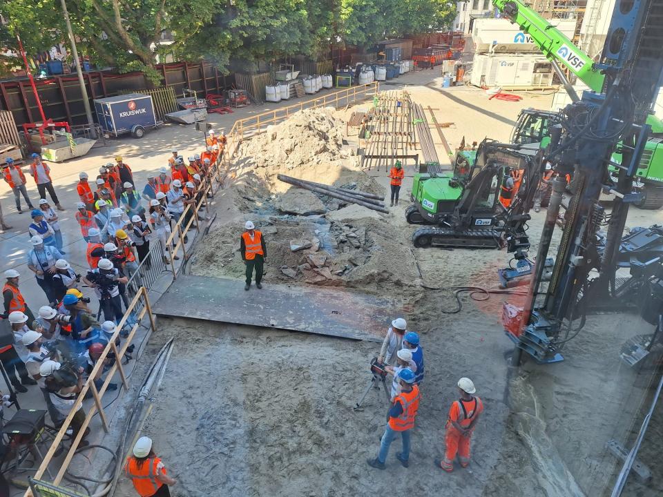 Beeld van boven van de bouwplaats: rechts grote machines, in het midden zand en materiaal en links mensen met bouwhelmen en veiligheidshesjes. 