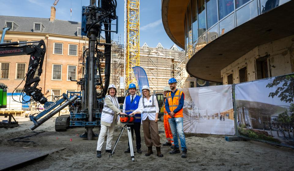 Vijf mensen staan buiten op een bouwplaats naast de plenaire zaal van de Tweede Kamer aan het Binnenhof. Kamervoorzitter Thom van Campen, tweede van links, heeft zijn hand op een knop die de boring in gang zet. 
