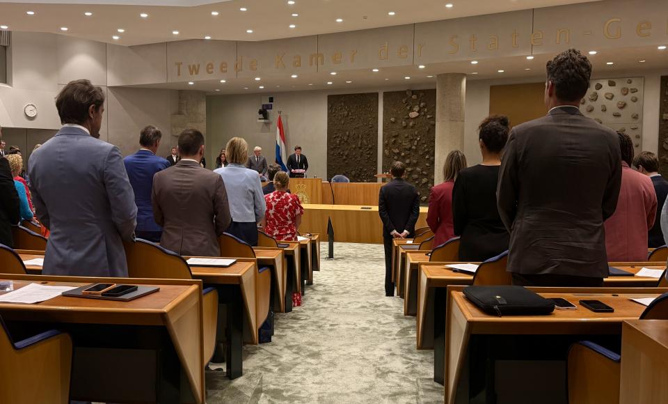 Kamerleden staan in de plenaire zaal bij een herdenking. Zij zijn op de rug gefotografeerd. 