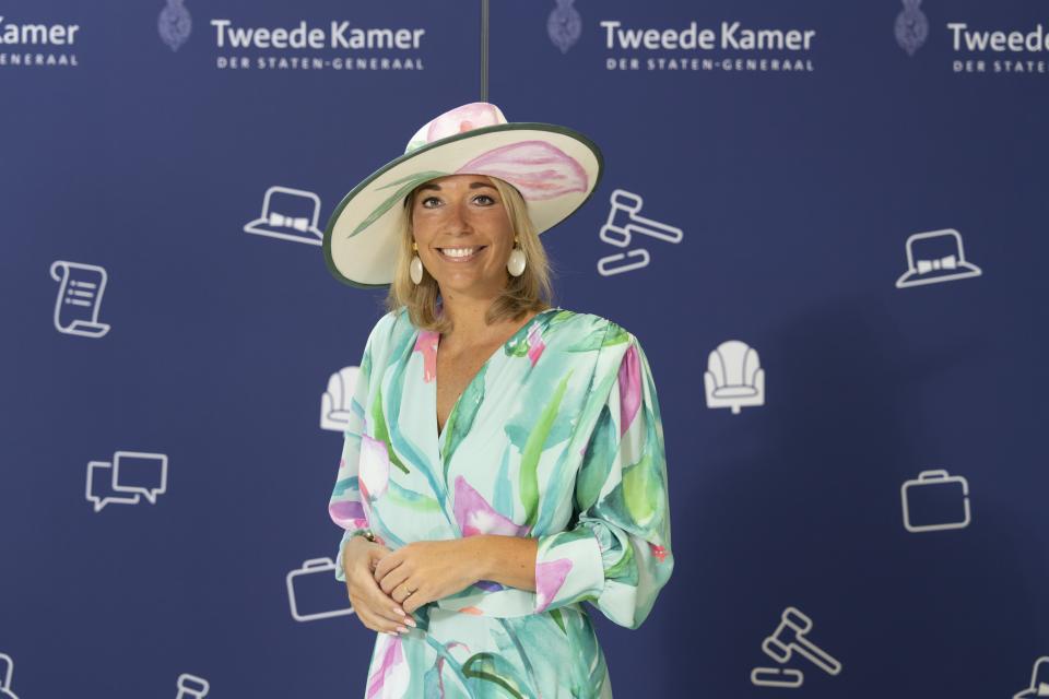 Kamerlid Ingrid Coenradie staat voor een blauwe wand met logo’s van de Tweede Kamer en afbeeldingen. Ze heeft een kleurige jurk aan en draagt een hoed.