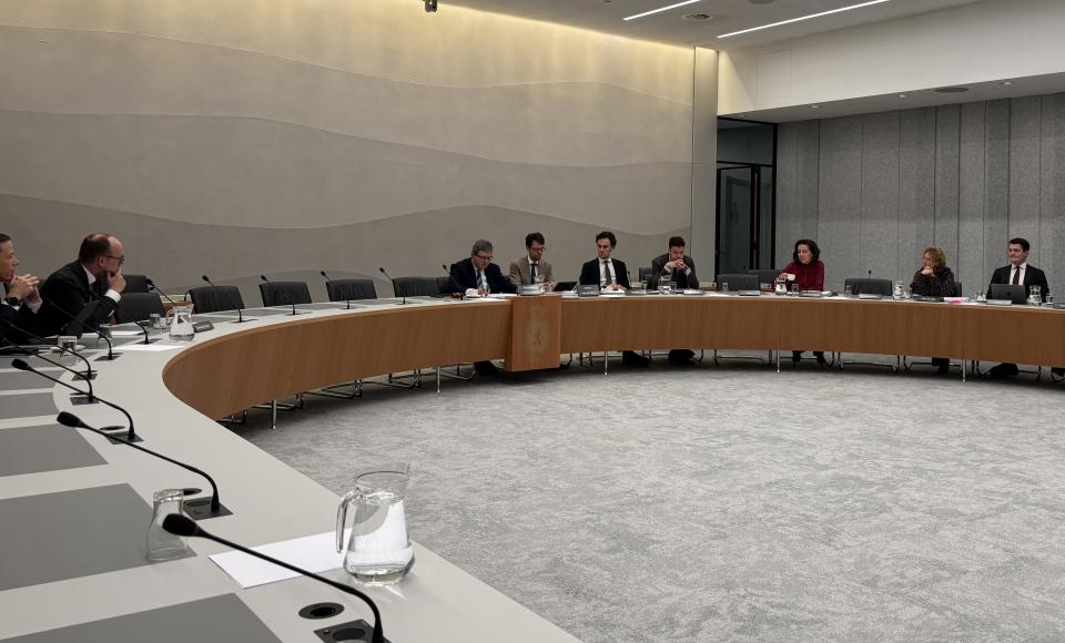 Aan een lange U-vormige tafel zit een aantal personen. Achter hen een grijze wand en op de voorgrond een grijs tapijt. 
