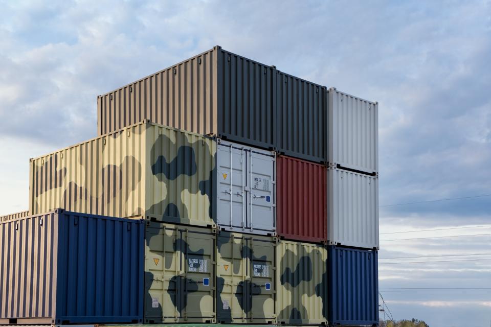Opgestapelde containers tegen een blauwe hemel, deels in camouflagekleuren.