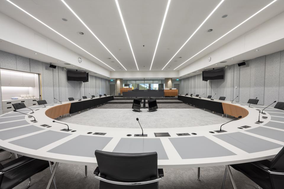 Beeld van lege zaal vanaf de halfronde tafel waaraan tijdens vergaderingen de Kamerleden en bewindspersonen zitten. In de verte is de publieke tribune zichtbaar en daarachter door het raam de Statenpassage. 