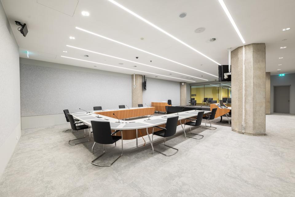 Overzicht van helverlichte witte zaal met in het midden een u-vormige opstelling van tafels met daarachter zwarte stoelen.