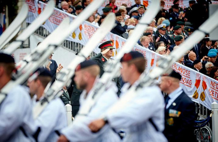 Veteranen marcheren in witte uniformen. Op de achtergrond staat publiek.