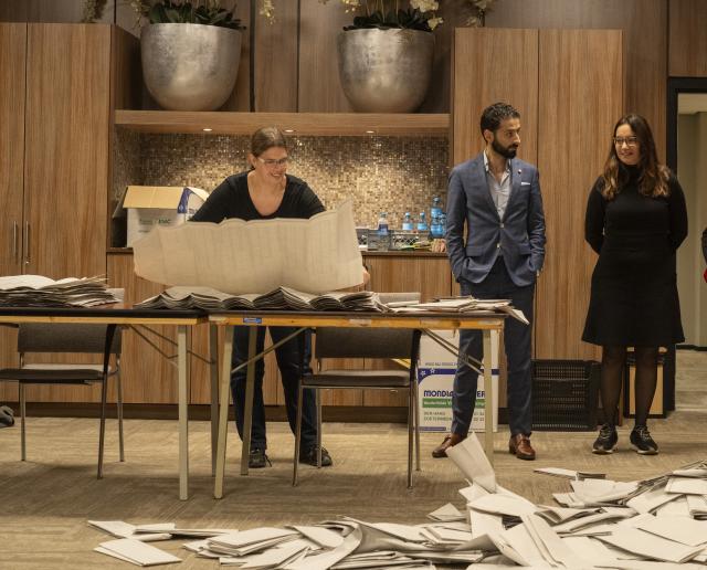 Staand achter een tafel vouwt een vrouw grote stembiljetten uit. Twee Kamerleden staan rechts achter haar en kijken toe.