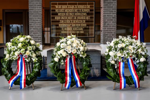 Op de foto staan drie kransen met witte bloemen. Ook is de Nederlandse vlag (rood, wit en blauw) te zien. 