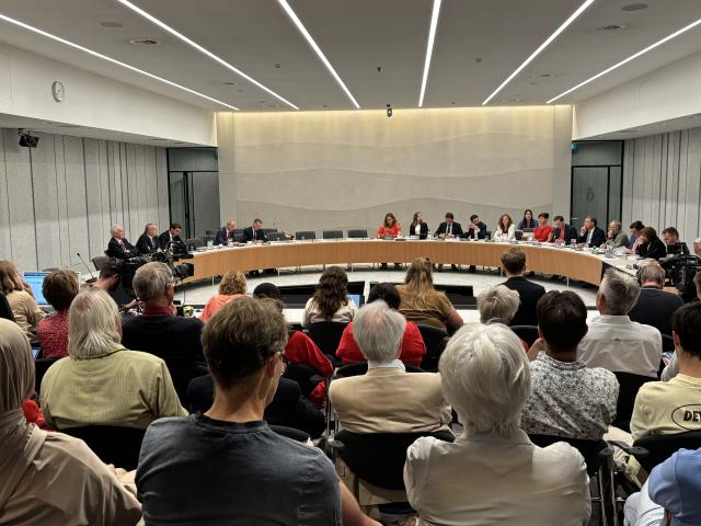 Commissiezaal met Kamerleden en publiek. Foto van achterin de zaal gemaakt, het publiek is van achteren te zien en de Kamerleden in de verte in een halve cirkel. 