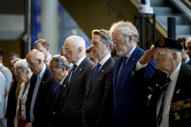Een rij aanwezigen staat, met gebogen hoofd, voor de zojuist gelegde kransen in de Statenpassage van de Tweede Kamer. 
