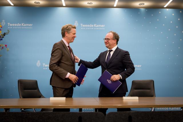 Twee mannen staan achter een tafel tegen een blauwe achterwand. Ze hebben allebei een boekwerk in de hand. 