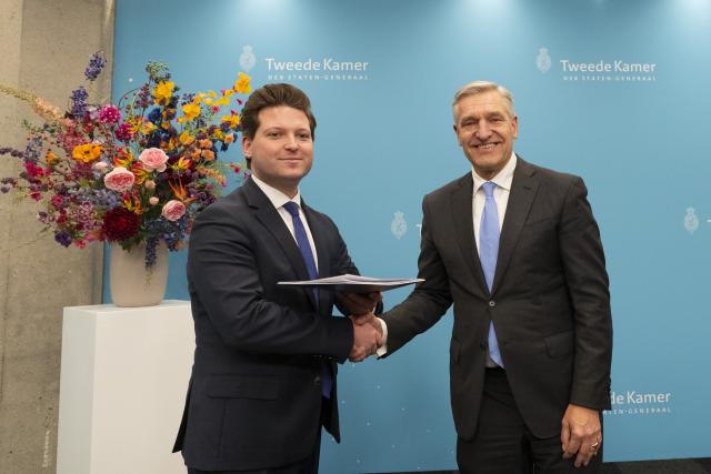 Kamervoorzitter Thom van Campen schudt de hand van informateur Sybrand Buma naast hem en houdt met de andere hand het verslag vast dat hij zojuist heeft gekregen. Beide mannen staan voor een lichtblauwe wand met het Kamerlogo, naast een bloemstuk op een pilaar. 