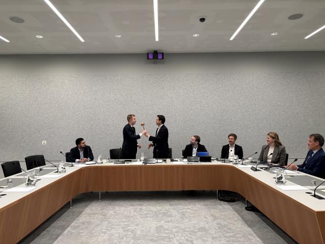 Kamerlid Julian Bushoff neemt de voorzittershamer over van collega Joost Sneller. Zij staan aan het uiteinde van een u-vormige rij tafels in een commissiezaal van de Tweede Kamer. Andere Kamerleden kijken toe vanachter de vergadertafel. 