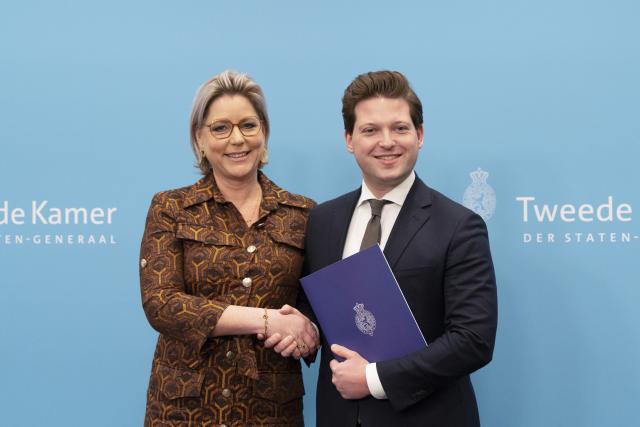 Twee personen staan naast elkaar voor een lichtblauwe wand met logo's van de Tweede Kamer. Links informateur Letschert, rechts Kamervoorzitter Thom van Campen. Zij schudden elkaar de hand en Van Campen houdt het eindverslag van Letschert voor zich. 