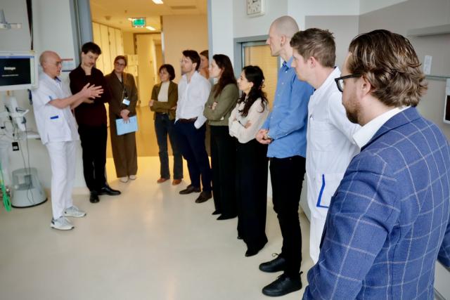 Leden en stafleden van de parlementaire enquêtecommissie Corona staan in een ziekenhuiskamer. Een arts spreekt hen toe.