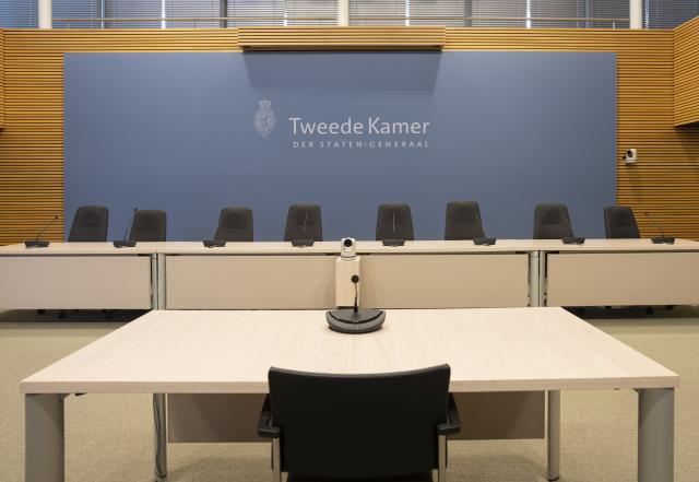 Zaal met op de voorgrond een stoel en tafel met microfoon. Op de achtergrond een tafel met daarachter acht stoelen en een scherm met het logo van de Tweede Kamer.