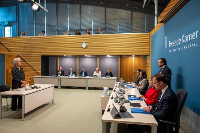 Overzichtsfoto van een verhoor in de Enquêtezaal. Getuige Marion Koopmans staat voor de commissieleden terwijl ze de belofte aflegt.