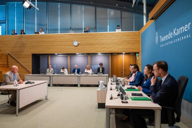 Rechts de leden van de commissie in een rij aan een lange tafel, van opzij gezien. Tegenover hen een getuige, alleen aan een tafel.  
