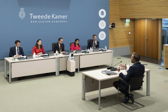 Getuige Hugo de Jonge, schuin van achteren in beeld, praat tegen de leden van de enquêtecommissie en gebaart daarbij met zijn handen. Tegenover hem zijn alle leden van de commissie in een rij zichtbaar. 