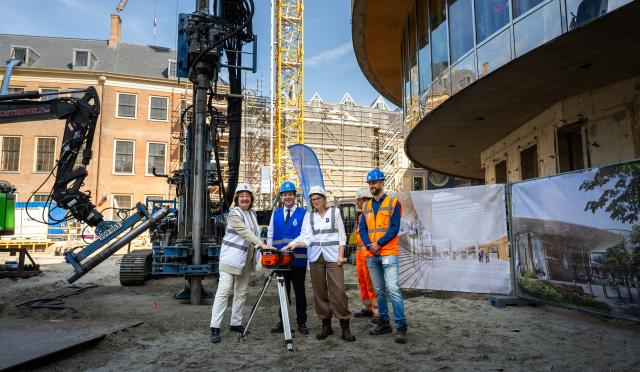 Vijf mensen staan buiten op een bouwplaats naast de plenaire zaal van de Tweede Kamer aan het Binnenhof. Kamervoorzitter Thom van Campen, tweede van links, heeft zijn hand op een knop die de boring in gang zet. 
