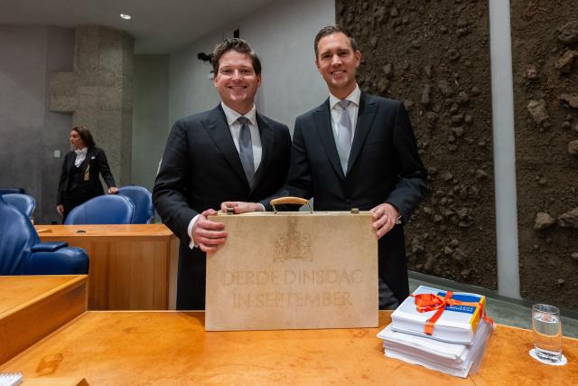 Kamervoorzitter Thom van Campen en minister van Financiën Eelco Heinen poseren samen staand in de plenaire zaal achter de ministerstafel. Ze houden allebei het koffertje met rijksbegroting en Miljoenennota vast. 