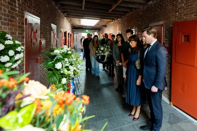 In de gang met gevangeniscellen staan de Voorzitters van Tweede en Eerste Kamer en andere aanwezigen in een lange rij. Zij kijken naar de deuren en de kransen een bloemstukken die neergezet zijn. 