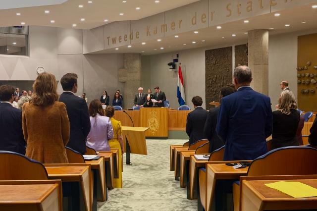 Kamerleden staan achter hun stoel in de plenaire zaal. Voorzitter Thom van Campen staat achter het spreekgestoelte en leest een toespraak voor.
