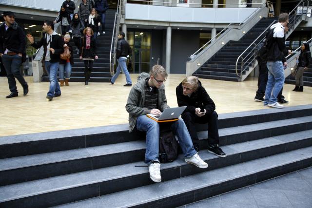 Twee studenten zitten op een trap van de aula van hun school. Een van hen heeft een laptop op zijn schoot en is daarop aan het werk. De ander zit ernaast en kijkt mee.