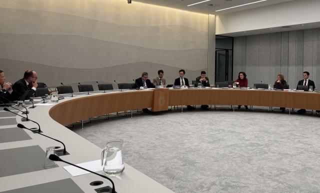 Aan een lange U-vormige tafel zit een aantal personen. Achter hen een grijze wand en op de voorgrond een grijs tapijt. 
