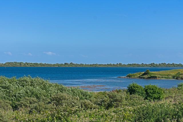 Weids uitzicht over het Grevelingenmeer, op de voorgrond groen grasland en daarachter helderblauw water en een blauwe lucht. 